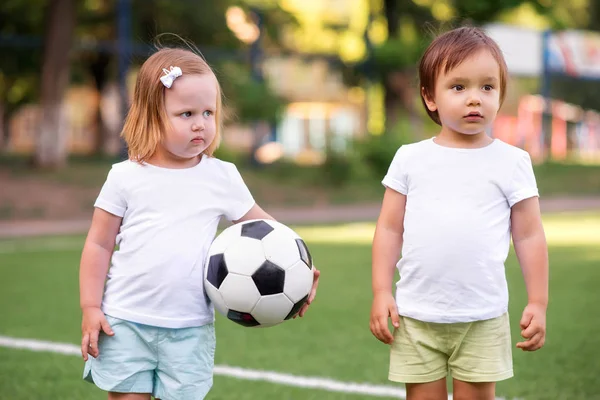 Equipo de fútbol: niña pequeña con pelota de fútbol y niño en uniforme deportivo de pie en el campo de fútbol y escuchando a alguien o algo. La chica está mirando al chico, el chico está mirando a un lado — Foto de Stock