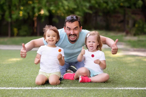 Familienspaß im Freien: Vater und zwei Kleinkinder, Junge und Mädchen, sitzen auf dem grünen Fußballplatz am Spielplatz und zeigen Daumen hoch! Kinder halten Bonbons auf Stöcken (Lutscher). glückliche gemeinsame Familienzeit — Stockfoto