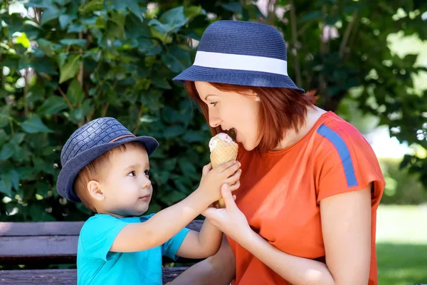Family leisure in park: mother and son in blue hats together sit on bench, boy is giving mom ice-cream in waffle cone to taste. Mom and child look at each other. Mother and son spend time together