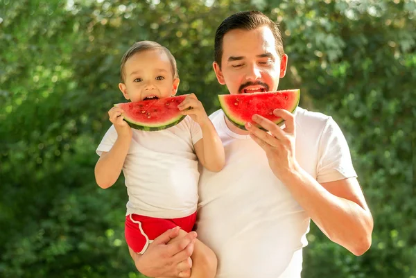 Portrait of dad and son simultaneously biting slices of watermelon outdoors. Father is holding child in arms. Green trees in background. Father and toddler son spending time together