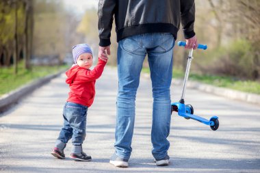 Baba ve küçük oğlu el ele tutuşarak yürüyüşe çıkıyorlar, arkadan bakın. Babam küçük mavi tekme scooter taşıyor, oğlu yarısı geri döndü ve geriye bakıyor
