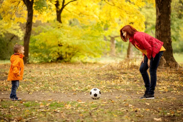Little boy playing football with mother in autumn park. Mom plays soccer with son outdoors: woman leaned forward explaining rules of game to child, ball is laying between them. Yellow trees in background. Active leisure and healthy parenting concept
