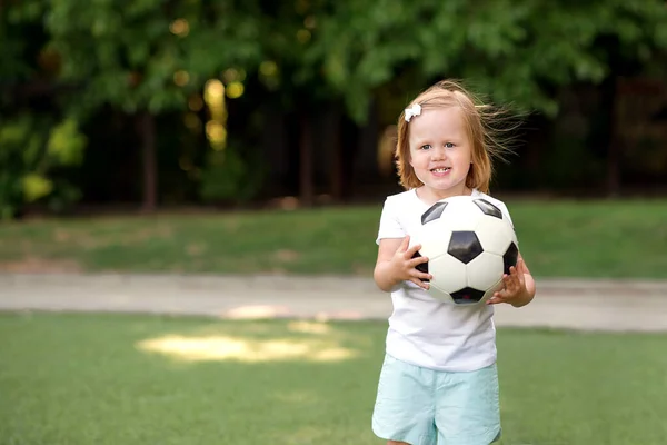 笑顔ブロンドの幼児の女の子は 夏の日に緑のサッカー場に立ってサッカーボールを保持しています スタジアムでボールで遊んでいる少女アスリートの肖像画 若いスポーツマン 女の子の力とアクティブな子供時代のコンセプト スペースのコピー — ストック写真