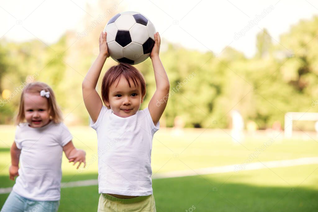 Little boy standing at football field holding soccer ball above head while smiling blonde girl is calmly sneaking from behind. Toddlers friends playing outdoors in sunny day. Copy space