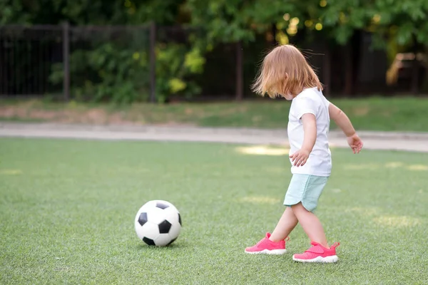 スタジアムでサッカーをしているピンクのスニーカーの幼児の女の子 小さなブロンドの子供は屋外の緑のフィールドでサッカーボールをキックする準備ができています 子供や将来のアスリートのコンセプトのための夏の活動 スペースのコピー — ストック写真