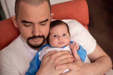 Baba kucağında bebek kızıyla koltukta uyuyor. Bebek uyandı ve şaşırmış bir suratla kameraya bakıyor. Yorgun baba küçük çocuğa bakıcılık yapıyor. Genç aileler uyku sorunları ve babalar günü konsepti. 