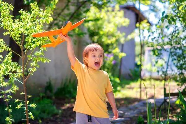 庭や中庭でオレンジ色のおもちゃの飛行機で一人で遊んで興奮した少年 幸せな幼児は屋外でおもちゃの航空機を進水させ 叫びなさい 将来のパイロットと積極的な子供時代のコンセプト スペースのコピー — ストック写真