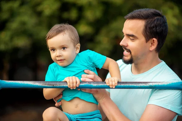 Father supporting toddler son doing muscle up exercise on steel bar. Dad and little son training together outdoors in summer evening. Healthy family, wellness and early development concept