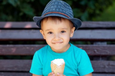 Mavi şapkalı küçük çocuk yaz ya da sonbahar günü waffle külahında dondurma yiyor. Gülümseyen çocuk parkta bankta oturmuş tatlı yiyor. Yaz hafta sonu ve tatil konsepti