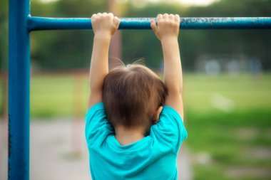 Küçük çocuk barfiks çekmeyi öğreniyor. Anaokulu öğrencisi spor sahasındaki yıpranmış çelik çubukta asılı, arkadan bakıyor. Azim, irade, çocuk ve spor anlayışı.