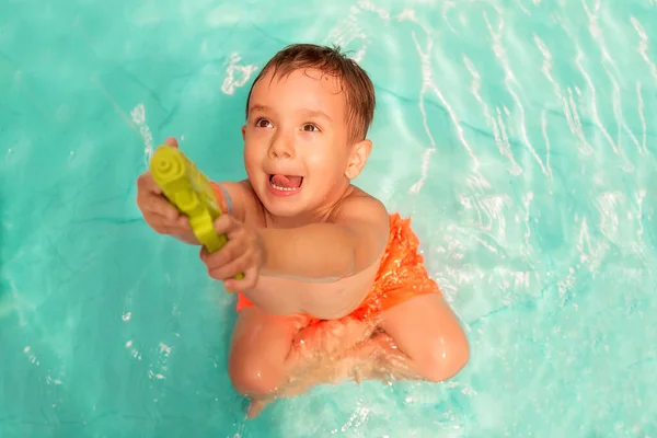 蹒跚学步的小男孩坐在游泳池里 脸上带着滑稽的表情 用绿色水枪射击 或瞄准 假期和户外活动对孩子的影响概念 — 图库照片