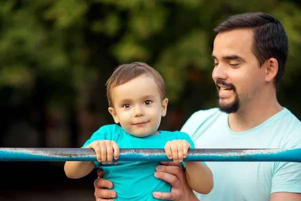 父は幼児の息子に屋外のスチールバーで運動を引き上げることを教えています 健康的な家族 子供のコンセプトのための親のサポートとスポーツ スペースのコピー ロイヤリティフリーのストック画像