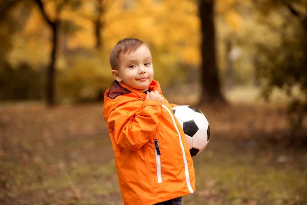 小さなサッカー選手 オレンジのジャケットの幼児の男の子は半分になって立ち 秋の公園や森の中でサッカーボールを保持します ボールを屋外で笑顔の子供 背景に黄色の紅葉 — ストック写真