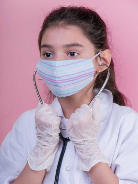 Doktor üniforması giyerken renkli steteskoplu komik bir kız. Renkli maske, panorama. çoklu uzay. Küçük bir doktor portresi. Kovid19. çözülebilir eldivenler 