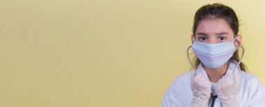 Doktor üniforması giyerken renkli steteskoplu komik bir kız. Renkli maske, panorama. çoklu uzay. Küçük bir doktor portresi. Kovid19. çözülebilir eldivenler 