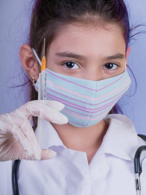 Doktor üniforması giyerken steteskopu ve şırıngası olan komik bir kız. Renkli maskeli Asyalı kız