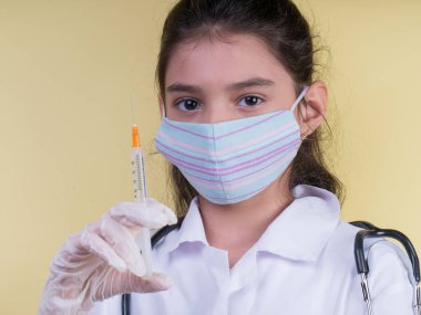 Doktor üniforması giyerken steteskopu ve şırıngası olan komik bir kız. Renkli maskeli Asyalı kız