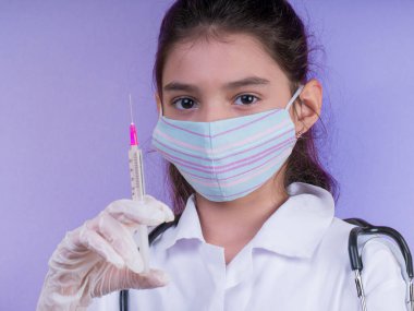 Doktor üniforması giyerken steteskopu ve şırıngası olan komik bir kız. Renkli maskeli Asyalı kız