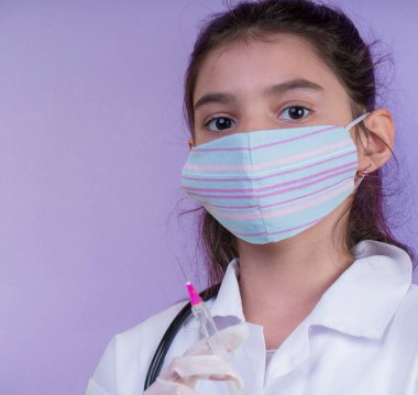 Doktor üniforması giyerken steteskopu ve şırıngası olan komik bir kız. Renkli maskeli Asyalı kız
