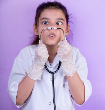 Doktor üniforması giyerken renkli steteskoplu komik bir kız. Heyecan ve cazibe konsepti. Coronavirus geçmişi. Aptal suratlar komik çılgın jestler. Komik ifade. 