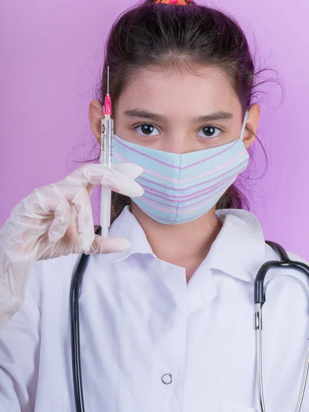 Doktor üniforması giyerken steteskopu ve şırıngası olan komik bir kız. Renkli maskeli Asyalı kız