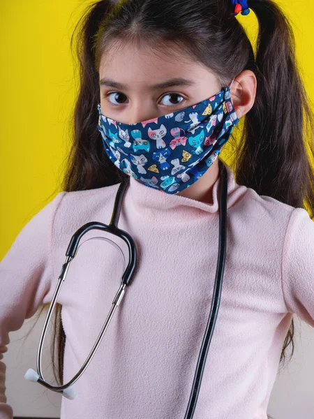 Sarı arka planda steteskop ve maske takan tatlı bir kız. Evde kal. Covid-19 'u durdurun. Virüs koruma ekipmanları. Karantinaya alın. Kız doktoru oynuyor. Eğitim ve işgal konsepti. Kendinde kal.