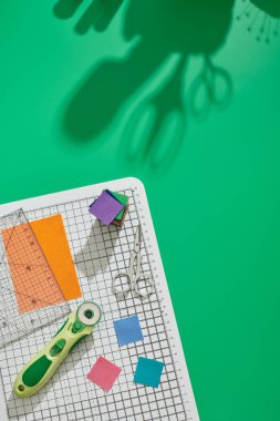 Döner kesici, cetvel, kumaş parlak kare parçaları, parlak kumaş kare parçalar yığını, zanaat mat üzerinde makas, yeşil arka plan