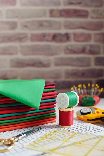 Stack di tessuti rossi e verdi circondati da accessori per cucito e trapuntatura su sfondo muro di mattoni — Foto Stock