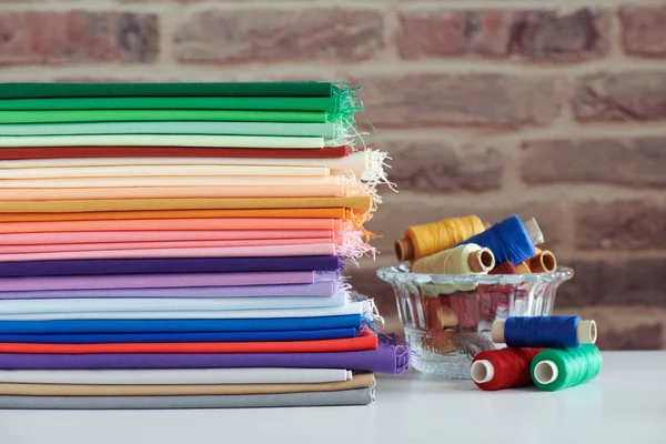 Stack of colorful monochromatic fabrics and spools of thread on brick wall background — Stock Photo, Image