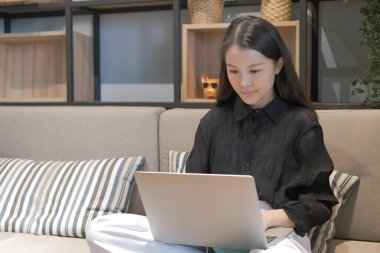 Evindeki koltukta oturan Asyalı ve beyaz dizüstü bilgisayar, evden çalışıyor. 