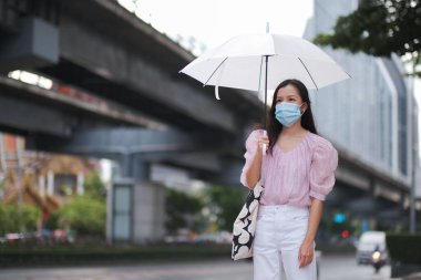 Cerrahi maske takan Asyalı kadın, yağmurlu bir günde COVID-19 'u önlemek için şemsiye tutuyor.