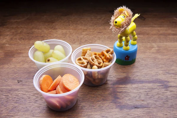 School lunch box snacks for kids over wooden background. Back to school. Healthy and fun snacks. Cute food art creative concepts. Toy, bowls with fruits, pretzels and vegetables.