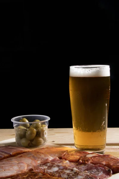 Soğuk bir bardak bira, zeytinli, jambonlu ve salamlı bir porsiyon aperatifli siyah arka planda ahşap masa. Mutlu saat zamanı..