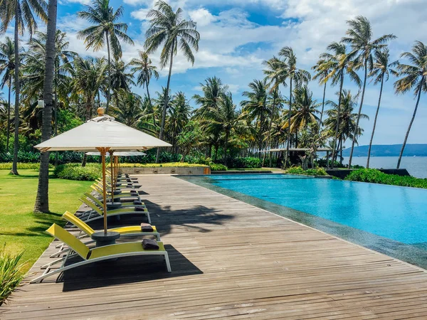 Beautiful tropical swimming pool in hotel or resort with umbrella, coconuts tree sun-loungers, palm trees with infinity pool view, ocean and mountain background