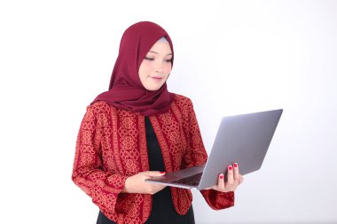 Genç Asyalı İslam kadını elinde dizüstü bilgisayarla ayakta ve gülümsüyor.