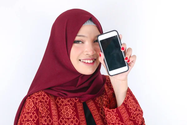 若いアジアのイスラム教徒の女性が笑顔で白い背景に立ってスマートフォンを示しています — ストック写真