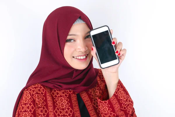 若いアジアのイスラム教徒の女性が笑顔で白い背景に立ってスマートフォンを示しています — ストック写真