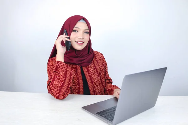 Young Asian Islam woman wearing headscarf is smiling and excited with when she call on the phone and front on the laptop on the table.