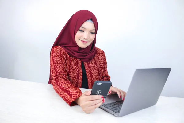 Young Asian Islam woman wearing headscarf is smiling on a mobile phone with laptop on the table.