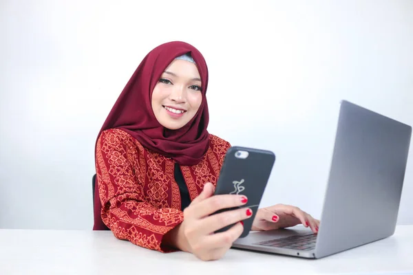 Young Asian Islam woman wearing headscarf is smiling on a mobile phone with laptop on the table.