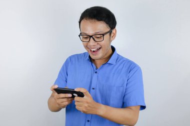 Asyalı genç adam oyun oynarken akıllı telefondan eğlenir ve gülümser. Endonezyalı adam mavi gömlek giyiyor..