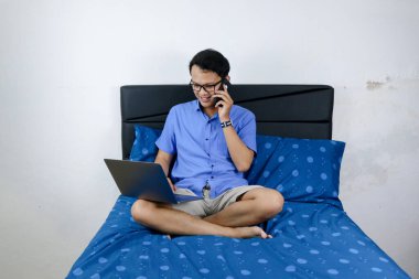 Genç Asyalı adam evde dizüstü bilgisayar ve telefonla çalışırken gülümsüyor ve eğleniyor. Yatakta oturup çalışan bir adam. Ev kavramından çalış.