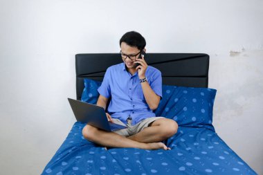 Genç Asyalı adam evde dizüstü bilgisayar ve telefonla çalışırken gülümsüyor ve eğleniyor. Yatakta oturup çalışan bir adam. Ev kavramından çalış.