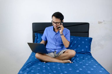 Genç Asyalı adam evde dizüstü bilgisayar ve telefonla çalışırken gülümsüyor ve eğleniyor. Yatakta oturup çalışan bir adam. Ev kavramından çalış.