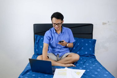 Genç Asyalı adam evde dizüstü bilgisayar ve telefonla çalışırken gülümsüyor ve eğleniyor. Yatakta oturup çalışan bir adam. Ev kavramından çalış.