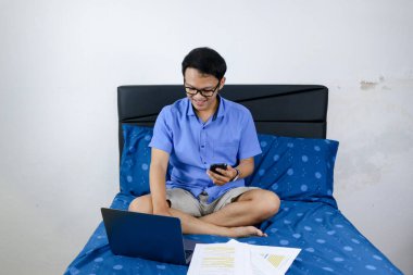 Genç Asyalı adam evde dizüstü bilgisayar ve telefonla çalışırken gülümsüyor ve eğleniyor. Yatakta oturup çalışan bir adam. Ev kavramından çalış.