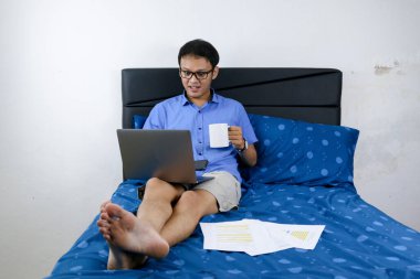 Genç Asyalı adam evde dizüstü bilgisayar ve bir fincan kahveyle çalışırken gülümsüyor ve eğleniyor. Yatakta oturup çalışan bir adam. Ev kavramından çalış.