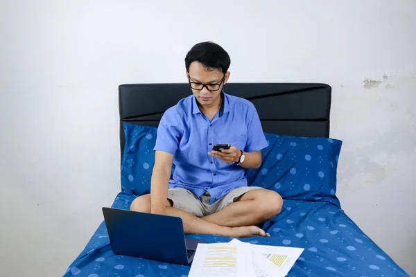 Young asian man is working at home with call in smartphone. A man sitting on bed and working. Work from home concept.