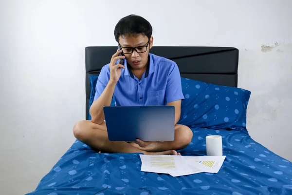 Young asian man is working at home with call in smartphone. A man sitting on bed and working. Work from home concept.