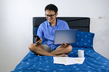 Genç Asyalı adam evde dizüstü bilgisayar ve telefonla çalışırken gülümsüyor ve eğleniyor. Yatakta oturup çalışan bir adam. Ev kavramından çalış.